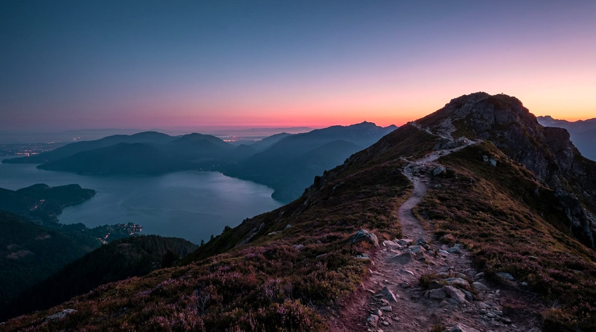 A mountain trail climbing a ridge above Howe Sound at dusk, the fjord and ranges below